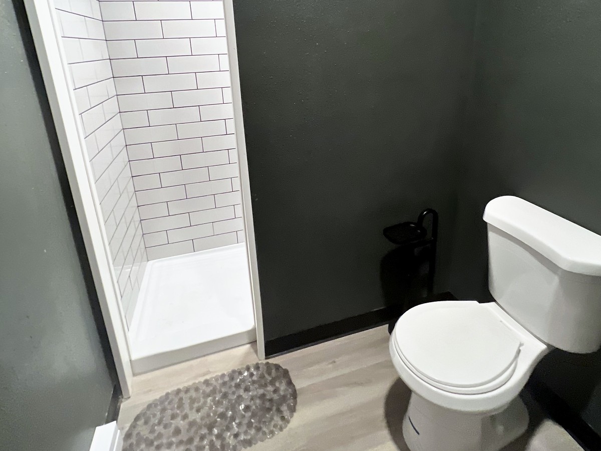 Inside the shared bathhouse — a white subway-tiled walk-in shower with a pebble bath mat and a flush toilet against deep charcoal-green walls