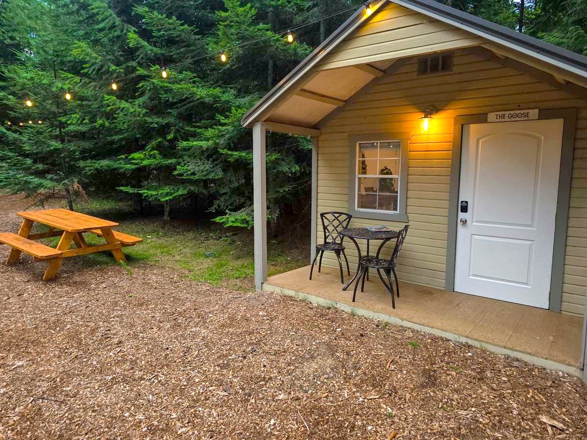 The Goose cabin at Round Mountain Retreat at dusk, the porch light on above a bistro table and a picnic table on the woodchips, string lights in the pines behind.