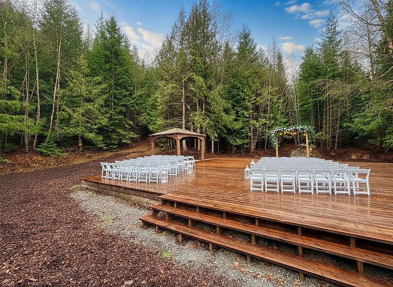 The open-air ceremony deck at Round Mountain Retreat set for a wedding — white chairs in rows either side of the aisle, a flower-dressed timber arbor at the treeline and evergreen forest all around.
