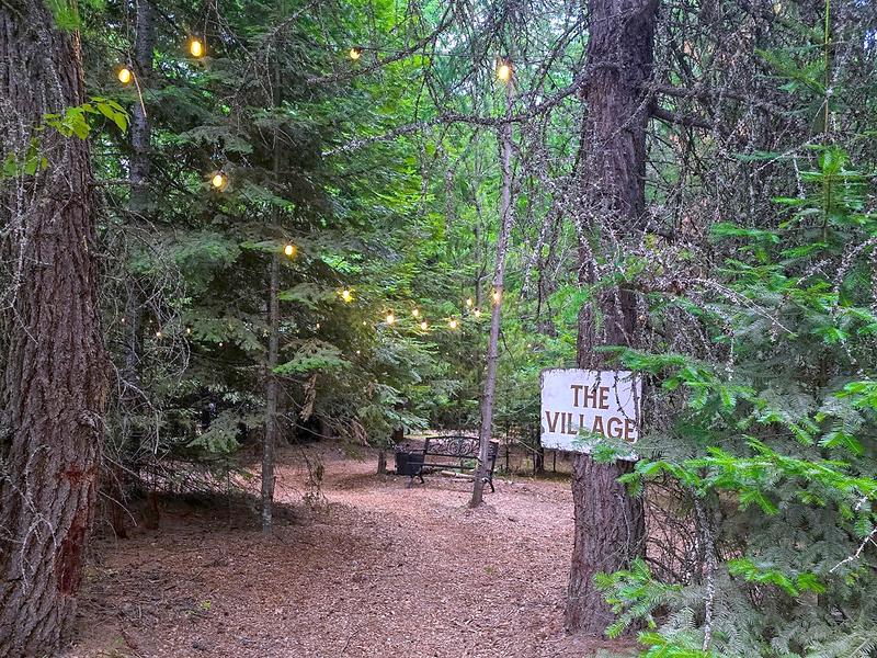 The soft woodchip path leading into The Village at Round Mountain Retreat, warm string lights glowing through the pines above a hand-painted sign reading THE VILLAGE.