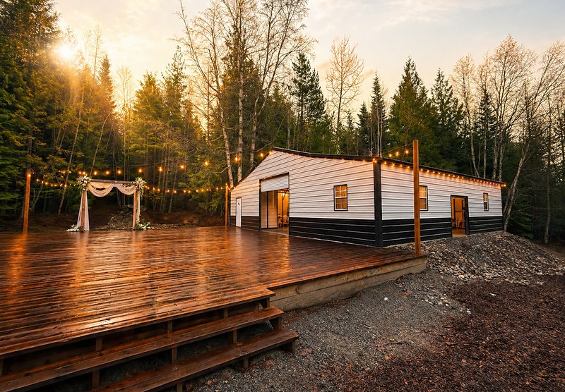 The event building at Round Mountain Retreat at golden hour, seen from the open-air deck — string lights strung across to the eaves, warm light in the rolled-up end door and the sun setting through the evergreens.