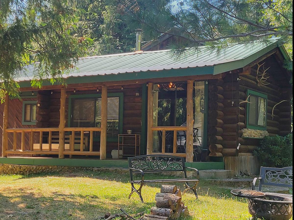 The Elk — a hand-built log cabin with a green metal roof and a covered porch on peeled-log posts, with iron benches, a fire bowl and split firewood on the lawn under the pines